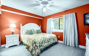 Ein Schlafzimmer mit einem Bett, einem Nachttisch, einer Lampe, einem Deckenventilator und einem Fenster mit Vorhängen.