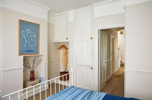 Un dormitorio con una cama, una mesita de noche con un jarrón y un libro, un perchero y una puerta que da a otra habitación.