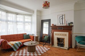 A living room with a sofa, a coffee table, a fireplace, and a large window with sheer curtains. - Elegant home in the heart of Wimbledon (London)