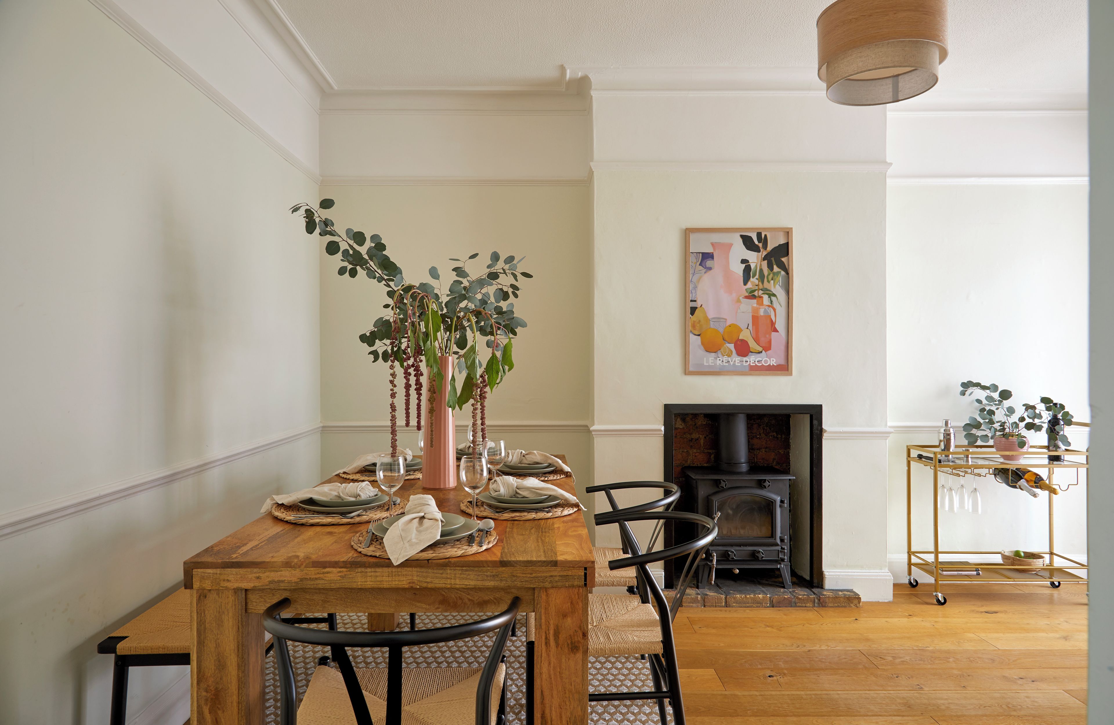 A dining room with a wooden table set for four, chairs, a vase with greenery, and a painting on the wall.