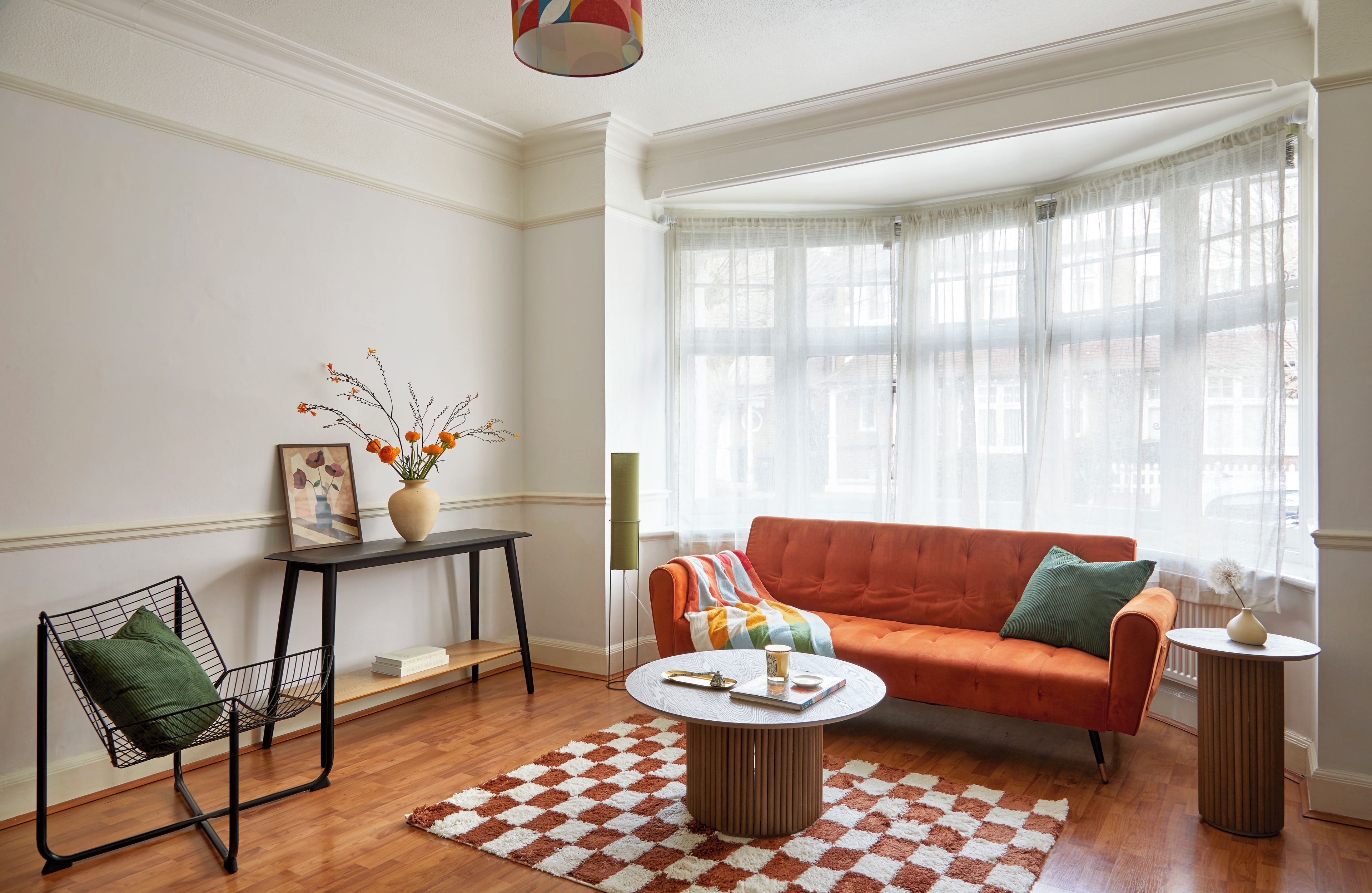 A living room with a sofa, coffee table, and a chair. There is a window with sheer curtains, a vase with flowers, and a framed picture on a console table.