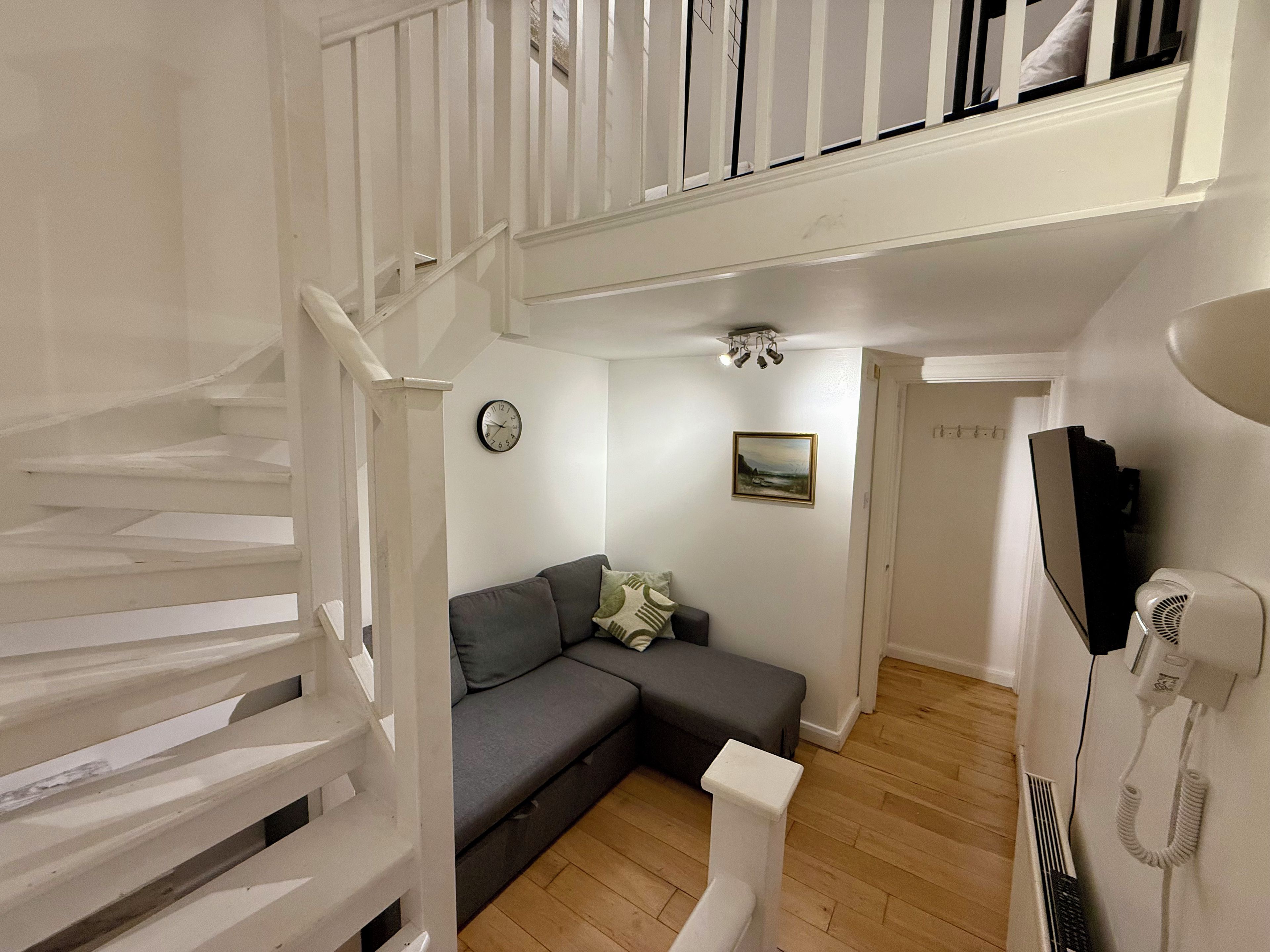 A staircase leading to a second floor, a grey sofa with cushions, a wall clock, a framed picture, and a telephone on the wall.
