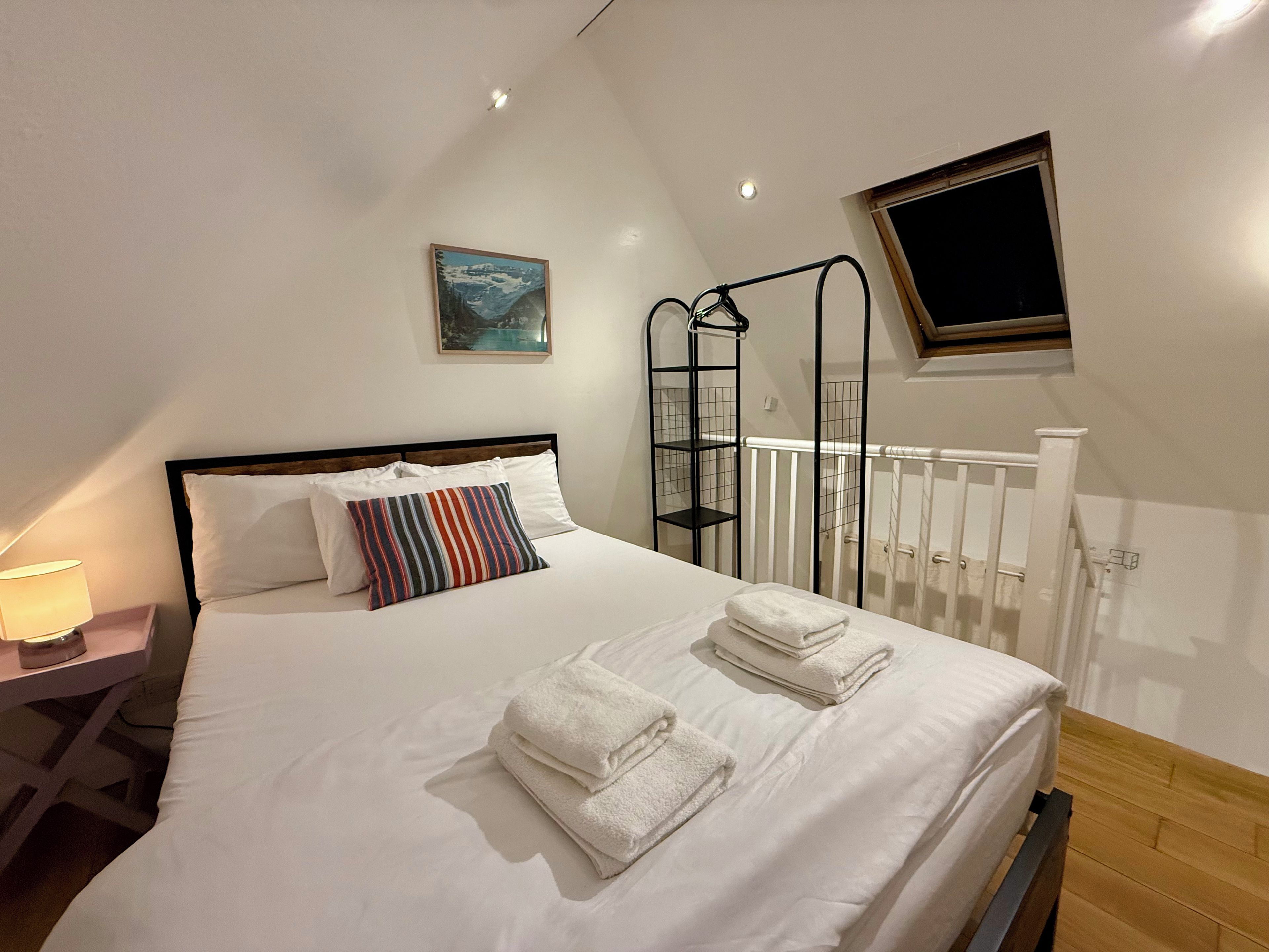 Un lit bien fait, avec du linge de lit blanc et des oreillers à rayures, une table de chevet avec une lampe, une fenêtre donnant sur un beau paysage et un tableau accroché au mur.