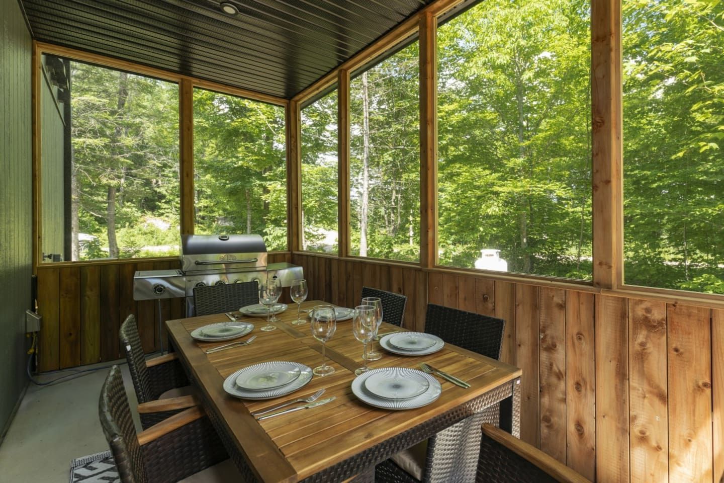 A covered outdoor dining area with a wooden table set for four, a grill, and a view of a forest.