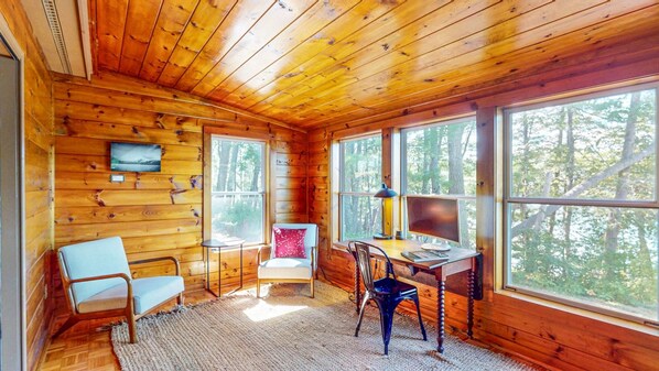 A cabin interior with wooden walls, a desk with a computer, two chairs, and large windows offering a forest view.