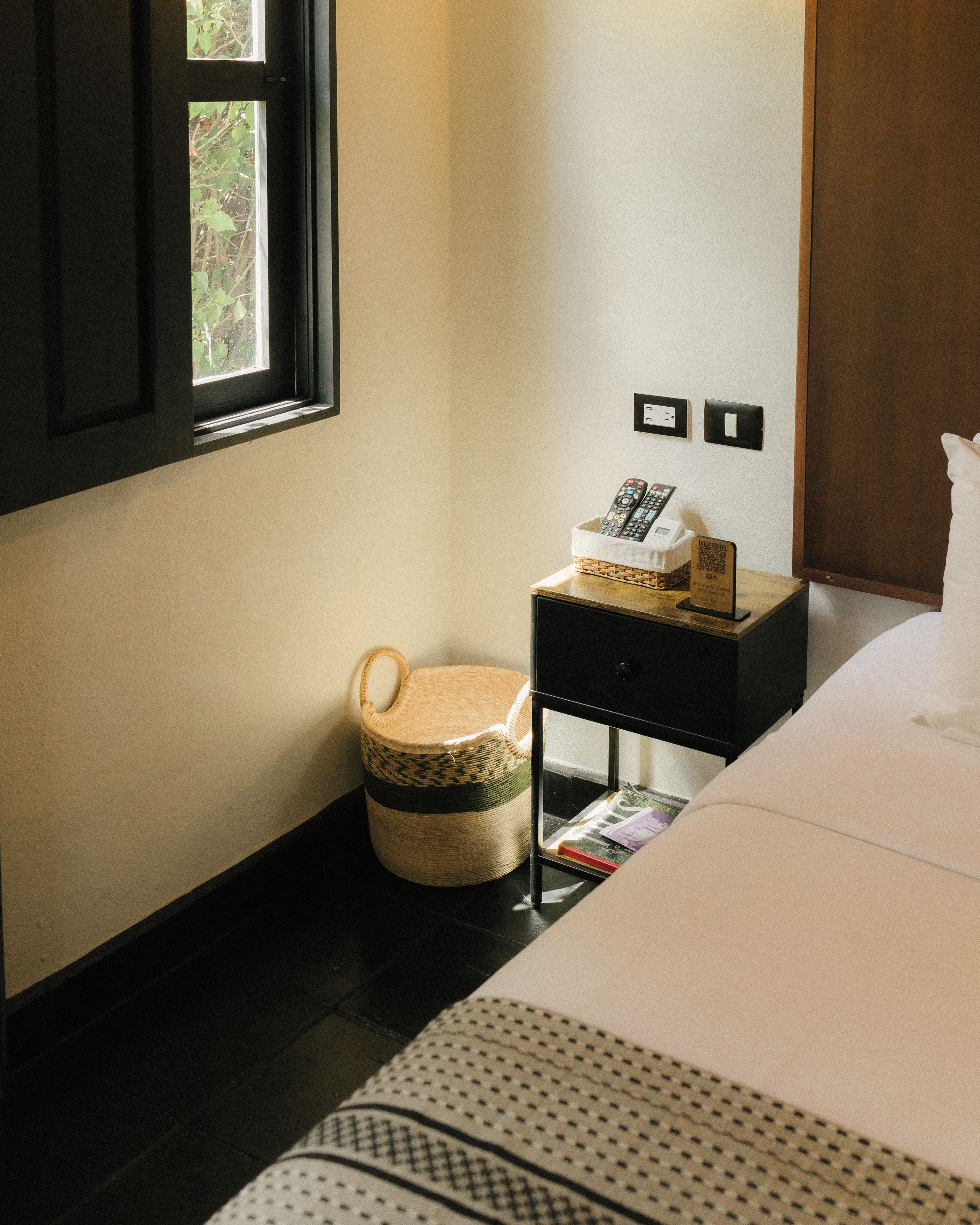 A hotel room with a bed, a small bedside table with a basket, a window, and a wall-mounted power outlet.