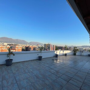 Una terrassa al terrat amb plantes en testos, cel blau clar i un paisatge urbà amb muntanyes al fons.