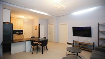 Ein modernes Wohnzimmer mit Flachbildfernseher, einem Essbereich mit Tisch und Stühlen sowie einer Küchenzeile mit Schränken und Kühlschrank.
