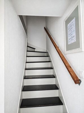 A staircase with black steps and a wooden handrail, leading to a landing with a white wall and a framed notice. - PLAGE ET GARE A PIED-MAISON TYPE LOFT- (Les Sables-d'Olonne)
