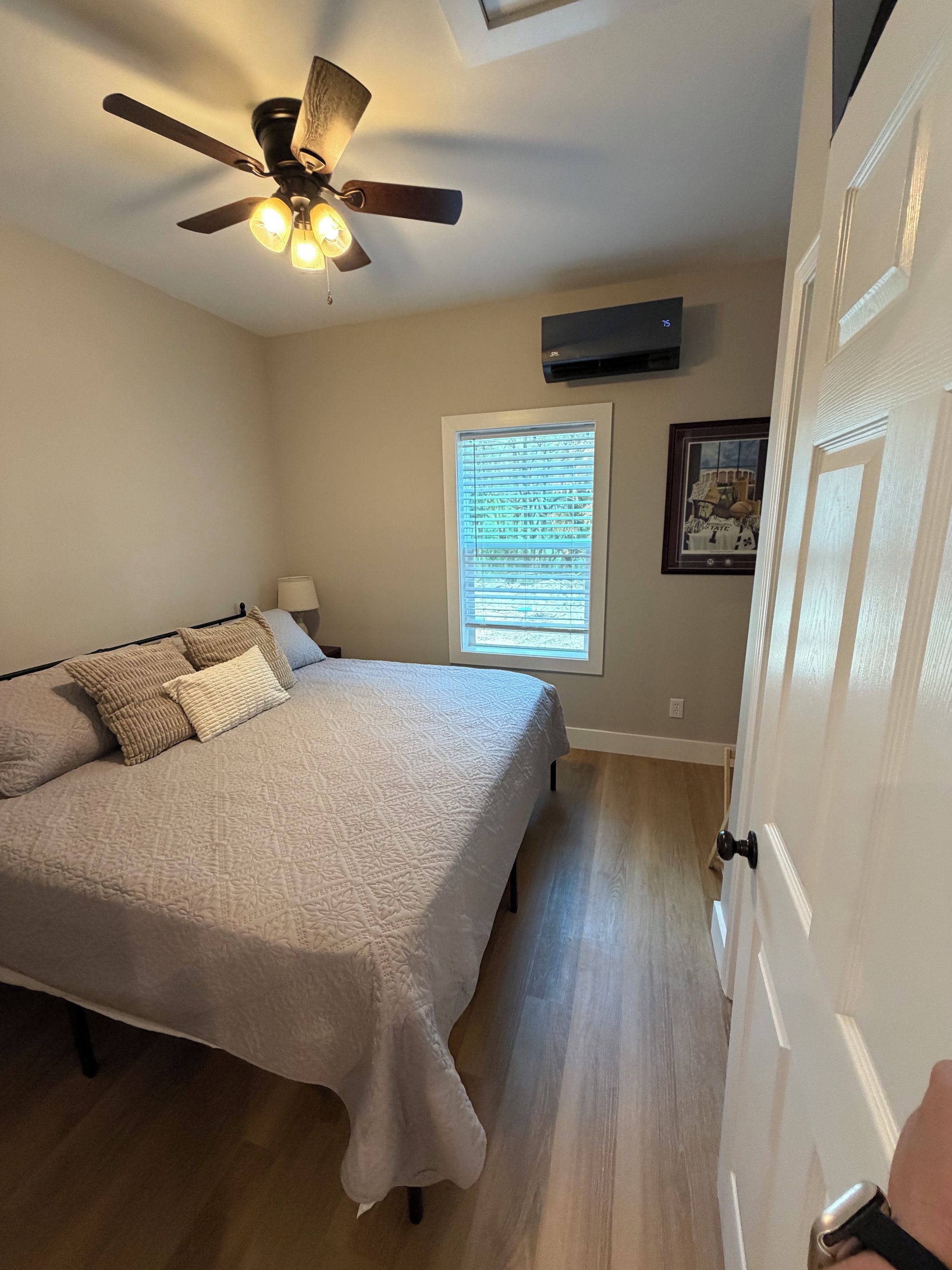 Une chambre à coucher comprenant un lit, un ventilateur de plafond, une fenêtre avec des stores, un climatiseur fixé au mur et une porte.