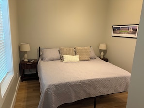 A bedroom with a bed, two bedside tables with lamps, a window with blinds, and a framed picture on the wall.