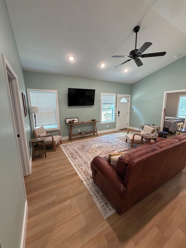A spacious living room with a large brown leather couch, a flat-screen TV mounted on the wall, and a ceiling fan.