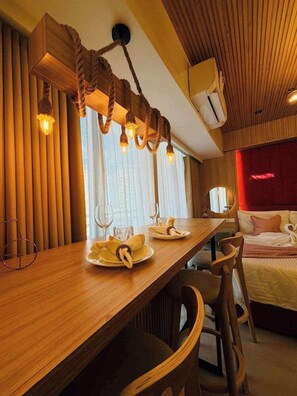 A hotel room with a wooden dining table, chairs, a bed with red headboard, and a hanging light fixture. - Studio Premium Suite (Paranaque)