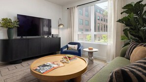 A modern living room with a flat-screen TV, a round coffee table with magazines, a blue armchair, and a view of buildings through large windows. - Inviting 1BR at Alba with Pool (Santa Clara)
