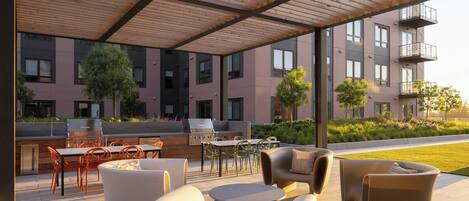 An outdoor patio with modern furniture, a barbecue grill, and a view of a multi-story building with balconies.