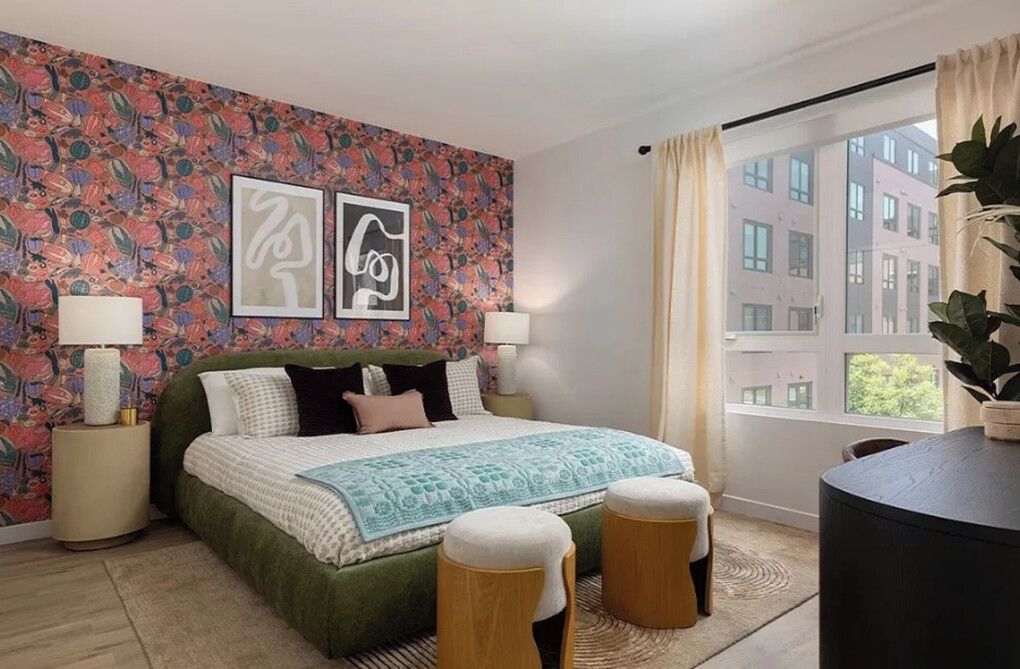 A bedroom with a bed, two bedside lamps, a patterned wall, a window with curtains, and a view of buildings outside.