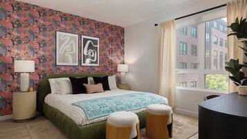 A bedroom with a bed, two bedside lamps, a patterned wall, a window with curtains, and a view of buildings outside.