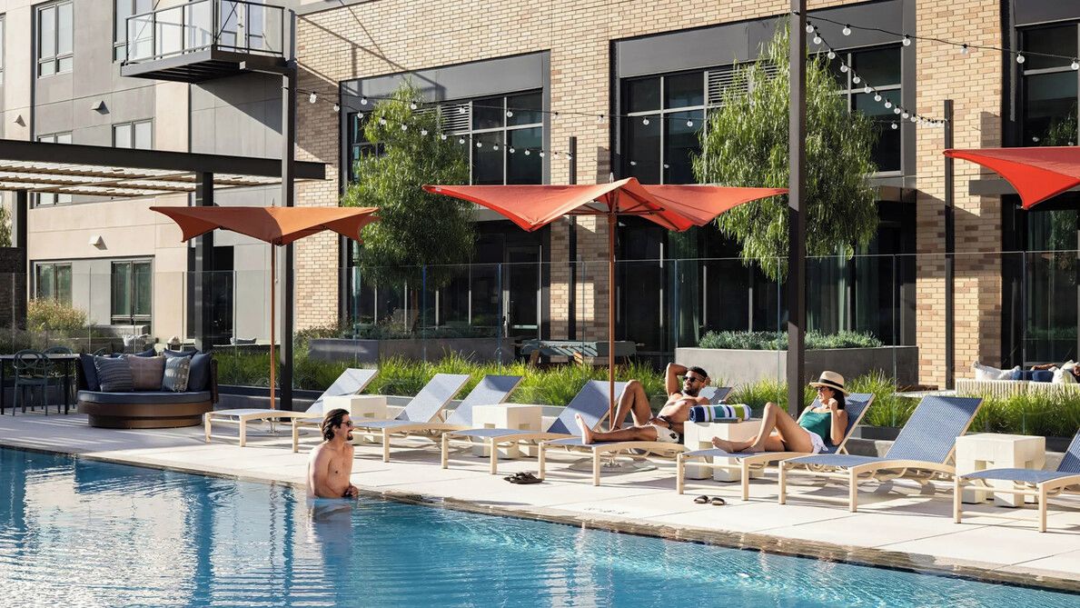 A poolside area with lounge chairs and umbrellas, a pool, and a building with large windows and a balcony.