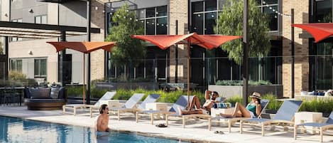 A poolside area with lounge chairs and umbrellas, a pool, and a building with large windows and a balcony.