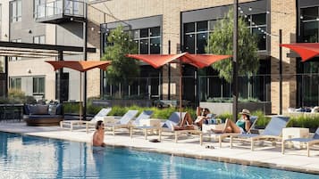 A poolside area with lounge chairs and umbrellas, a pool, and a building with large windows and a balcony.