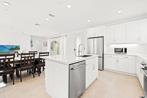 A modern kitchen with white cabinetry, stainless steel appliances, and a dining area with wooden chairs and a table. - Modern Desert Retreat 3BR Home w/ Community Pool (Palm Desert)