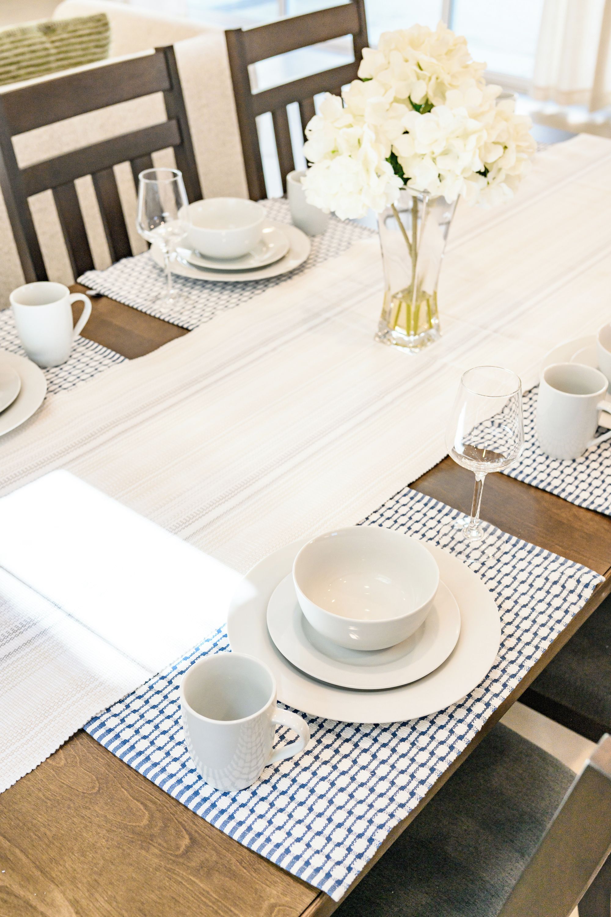 Una mesa de comedor dispuesta con platos, tazones y tazas blancos, un florero con flores blancas y un vaso de agua.