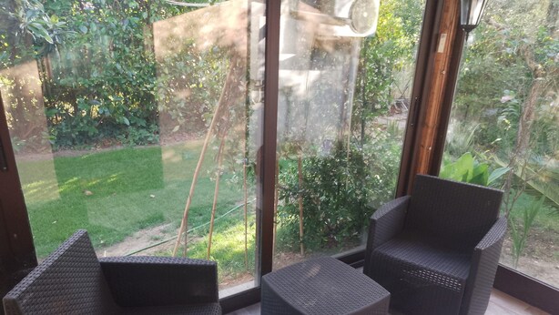 Una stanza con due poltrone in vimini scuro e un tavolino, ampie finestre con vista su un giardino curato.