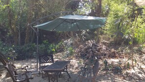 Area esterna con tavolo e due sedie sotto un ombrello verde, immersa tra alberi e vegetazione.