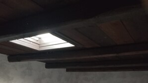 Una stanza piccola, scarsamente illuminata, con un letto singolo, una sedia rossa e un soffitto inclinato con una finestra a lucernario.