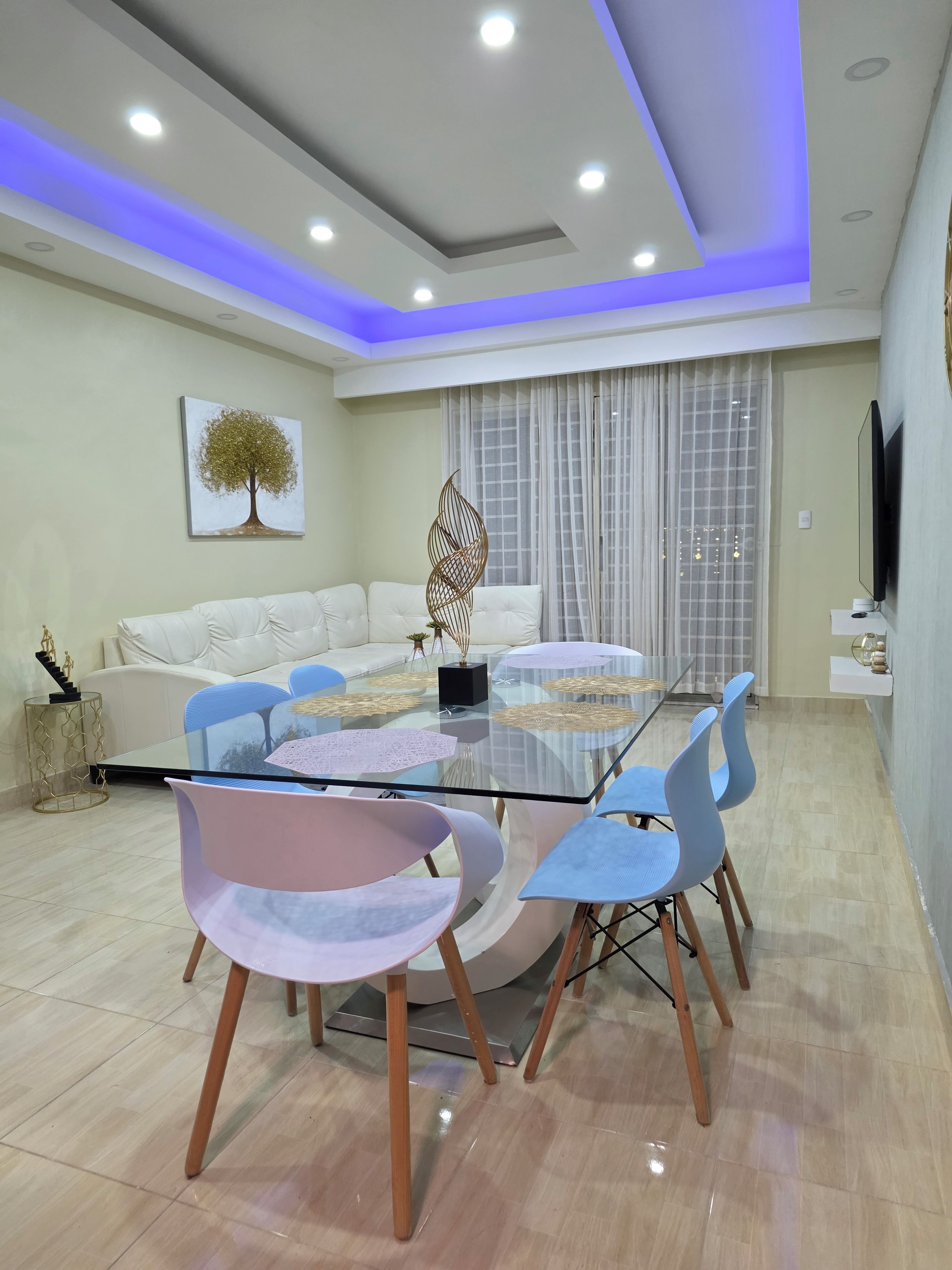 A modern dining area with a glass table, multiple chairs, a sofa, and a decorative tree painting on the wall.
