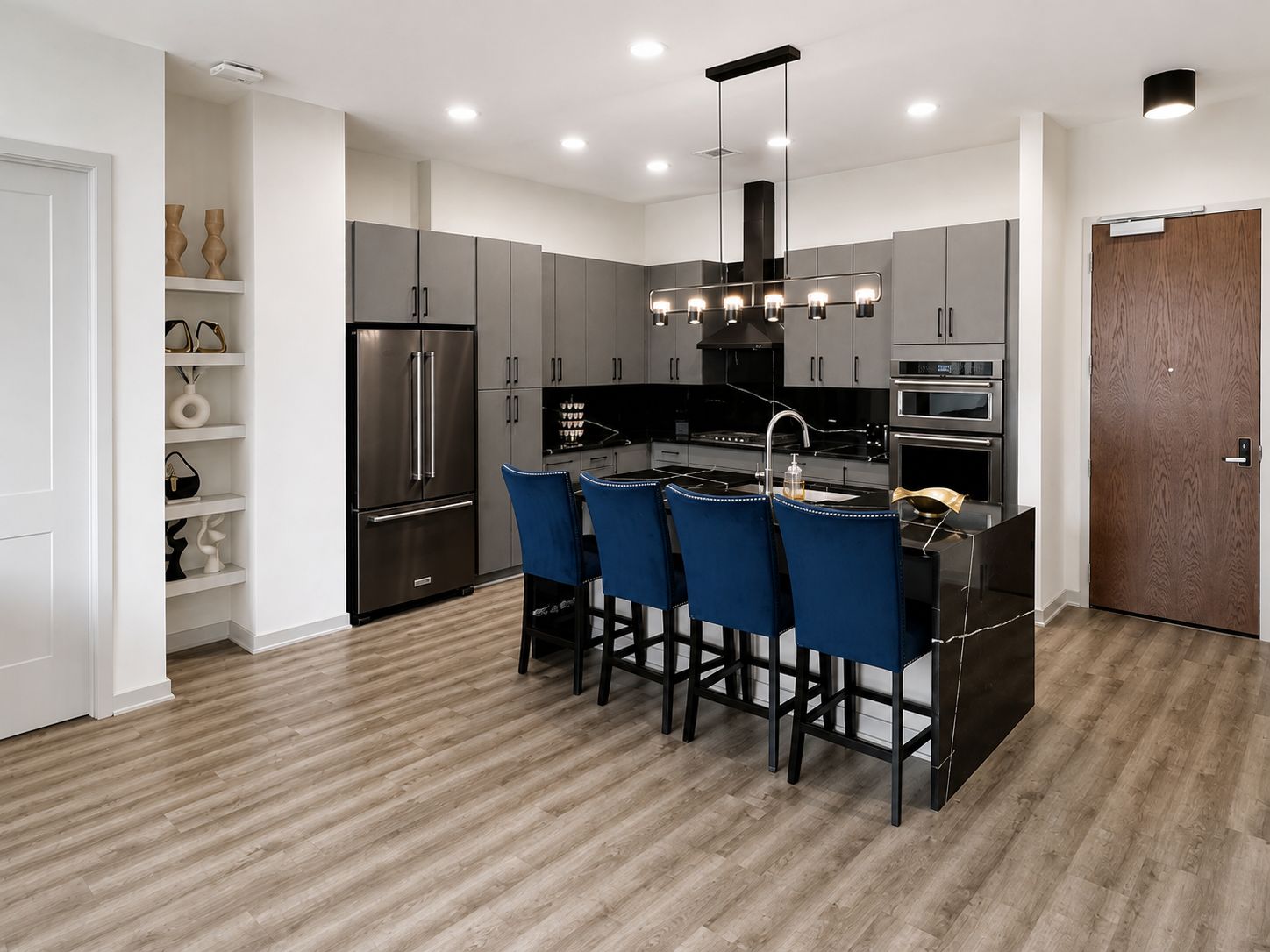 A modern kitchen with grey cabinets, a stainless steel refrigerator, a built-in oven, and a central island with blue bar stools.