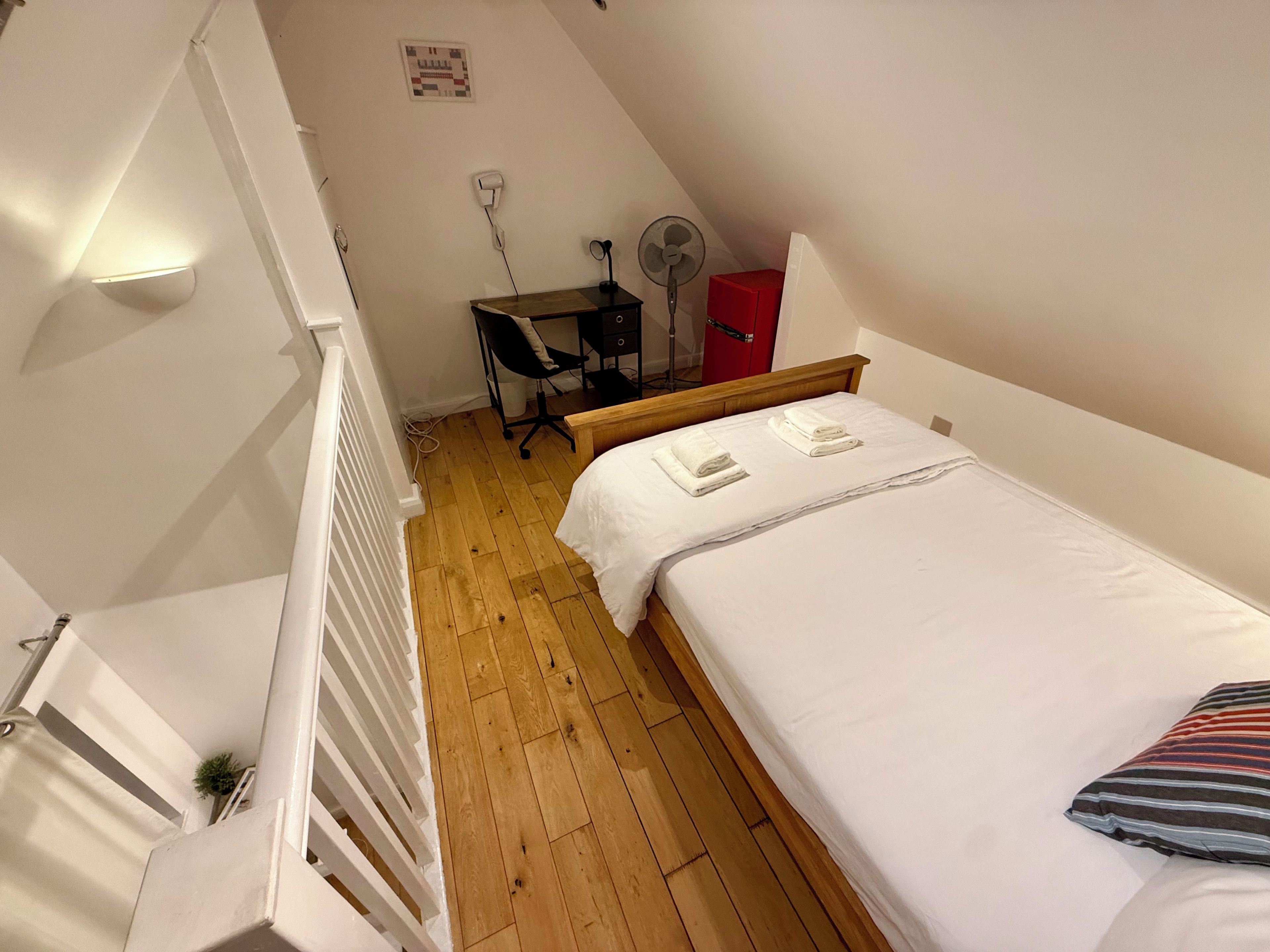 Een slaapkamer met een houten vloer, een bed met witte lakens, een bureau met stoel, een rode kast en een staande ventilator.