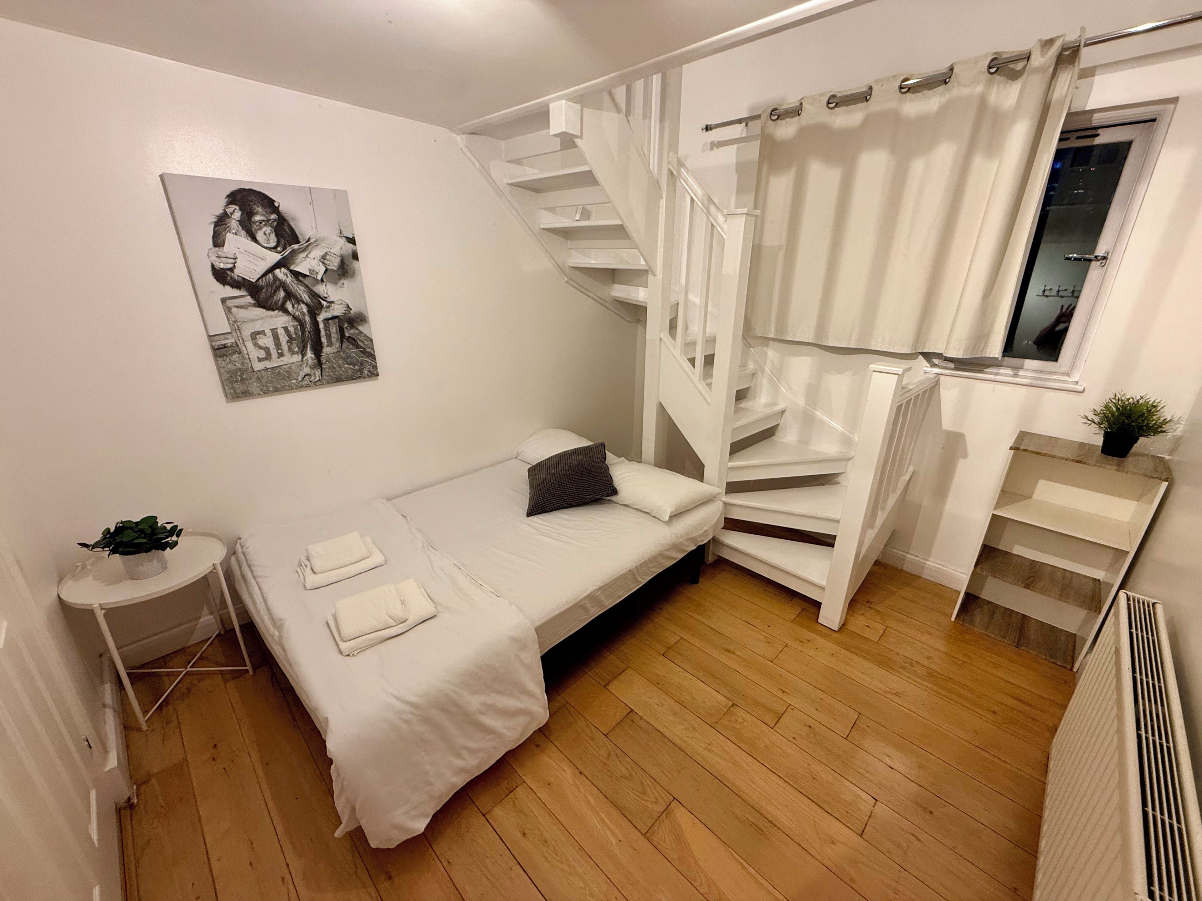A small, white-themed bedroom with a single bed, a white staircase, a small table with a plant, and a wall-mounted picture.