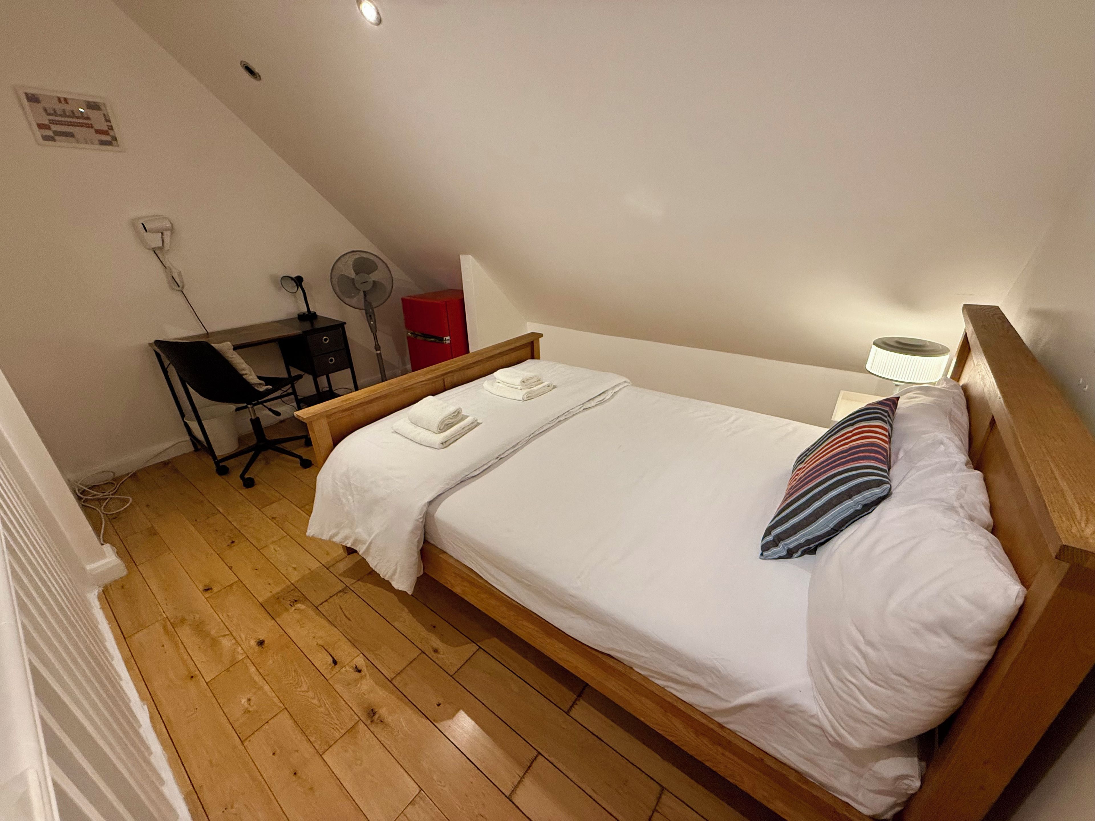 Un llit individual amb roba de llit blanca, un escriptori amb una cadira, un armari vermell, un ventilador de peu i una làmpada fixa a la paret.