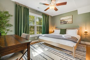 Ein Schlafzimmer mit Bett, Schreibtisch, Stuhl, Deckenventilator, einem Fenster mit Vorhängen, einer Wandleuchte und einem Gemälde.