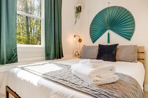 A neatly made bed with a white comforter and a folded white towel, a teal wall fan, a framed picture on the wall, and a window with green curtains. - Spacious w/ Game Room + Hot Tub | Mins to Downtown (Woodfin)
