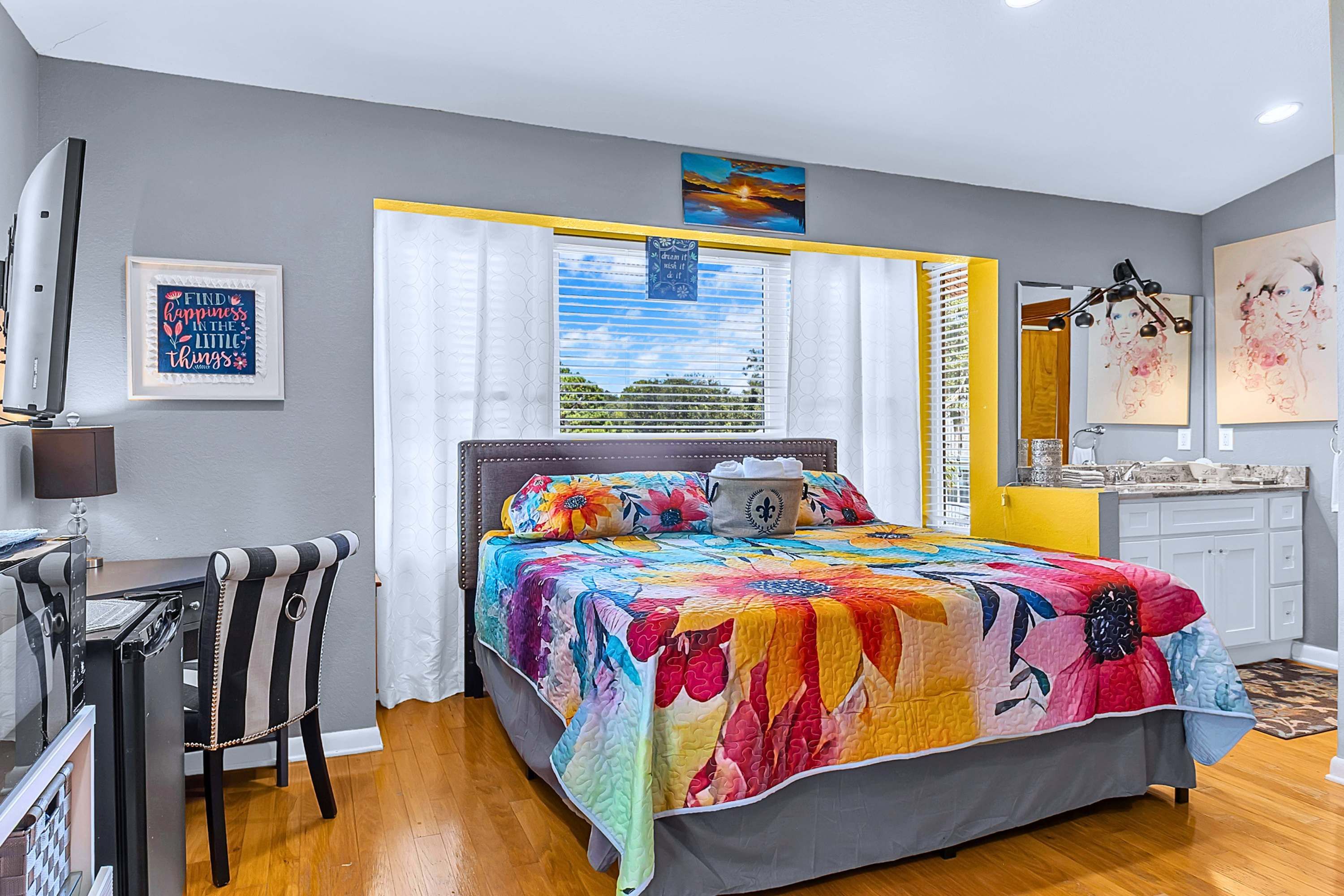 A bedroom with a bed, a desk, a chair, a window with curtains, and artwork on the walls.