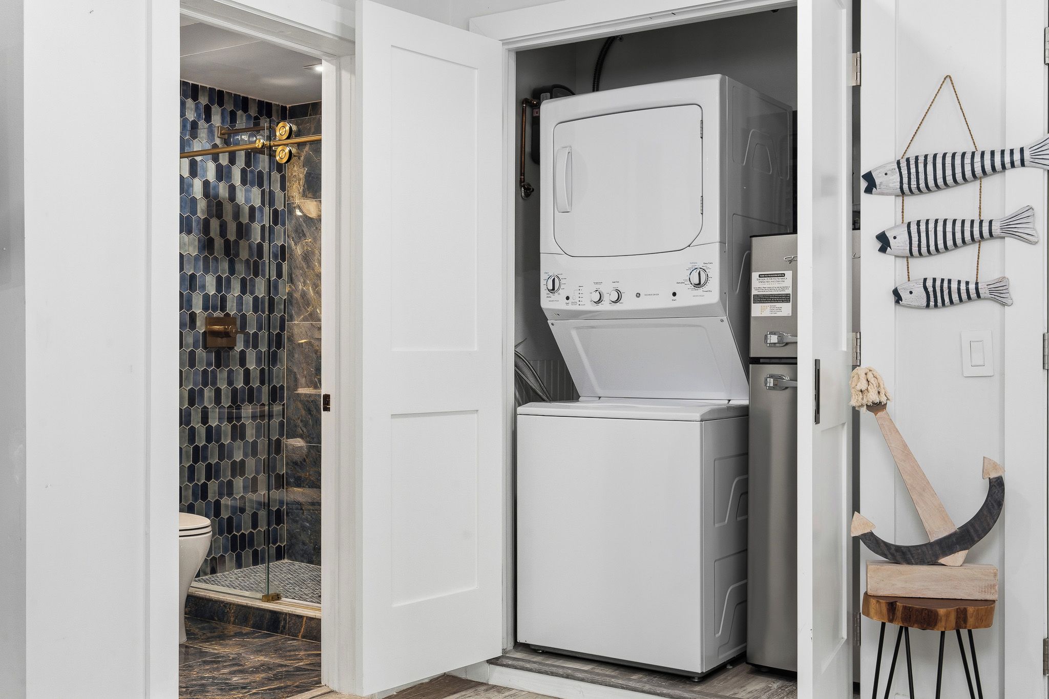 A compact laundry room with a washing machine and dryer, a tiled shower area, and a small wooden stool.