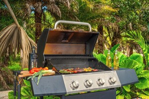 Een barbecue met vier branders, een fles kookolie en groenten op een houten plank.