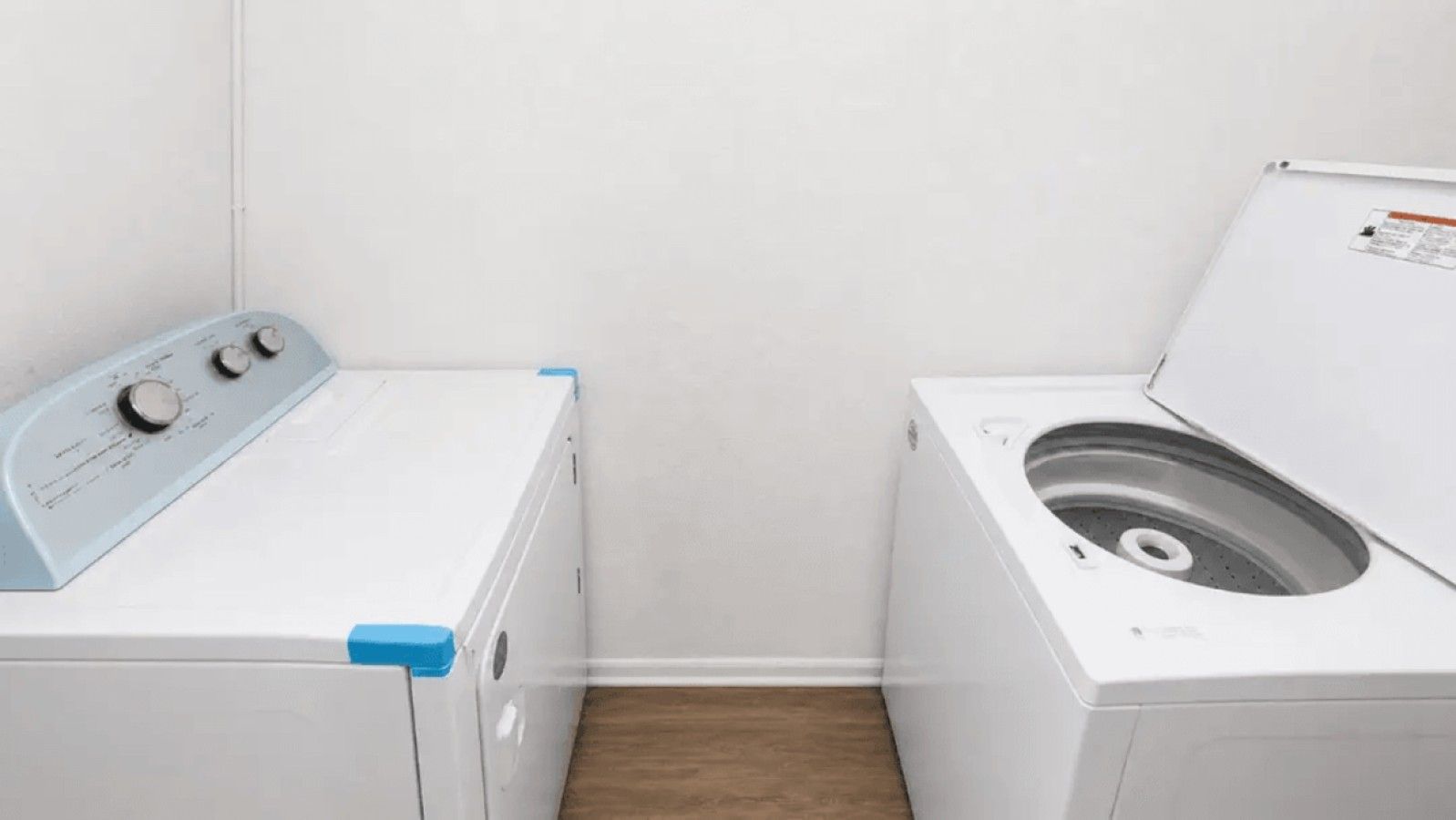 A laundry room with a front-loading washer and dryer, a wooden floor, and a white wall.