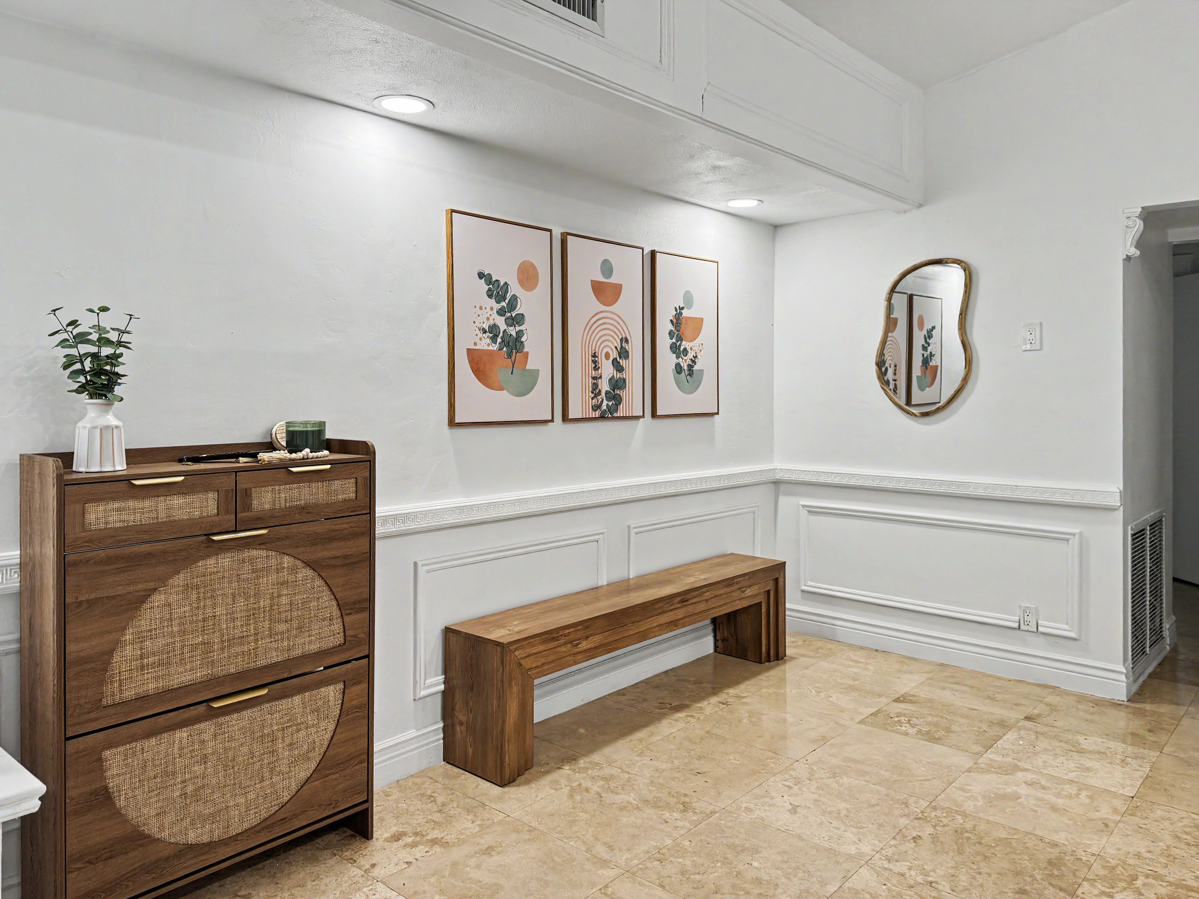 Un pasillo con un banco de madera, una pared blanca con tres obras de arte enmarcadas, una consola blanca con un florero y una planta, y un espejo en la pared.