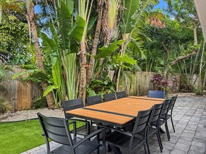 An outdoor patio with a wooden table and black chairs, surrounded by lush tropical plants and a wooden fence. - Ultimate Group Getaway – Pool, Games & Tiki Bar (Wilton Manors)