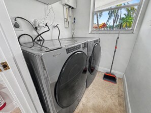 A laundry room with two Whirlpool washers, a window with a view of palm trees, and a mop leaning against the wall. - Ultimate Group Getaway – Pool, Games & Tiki Bar (Wilton Manors)