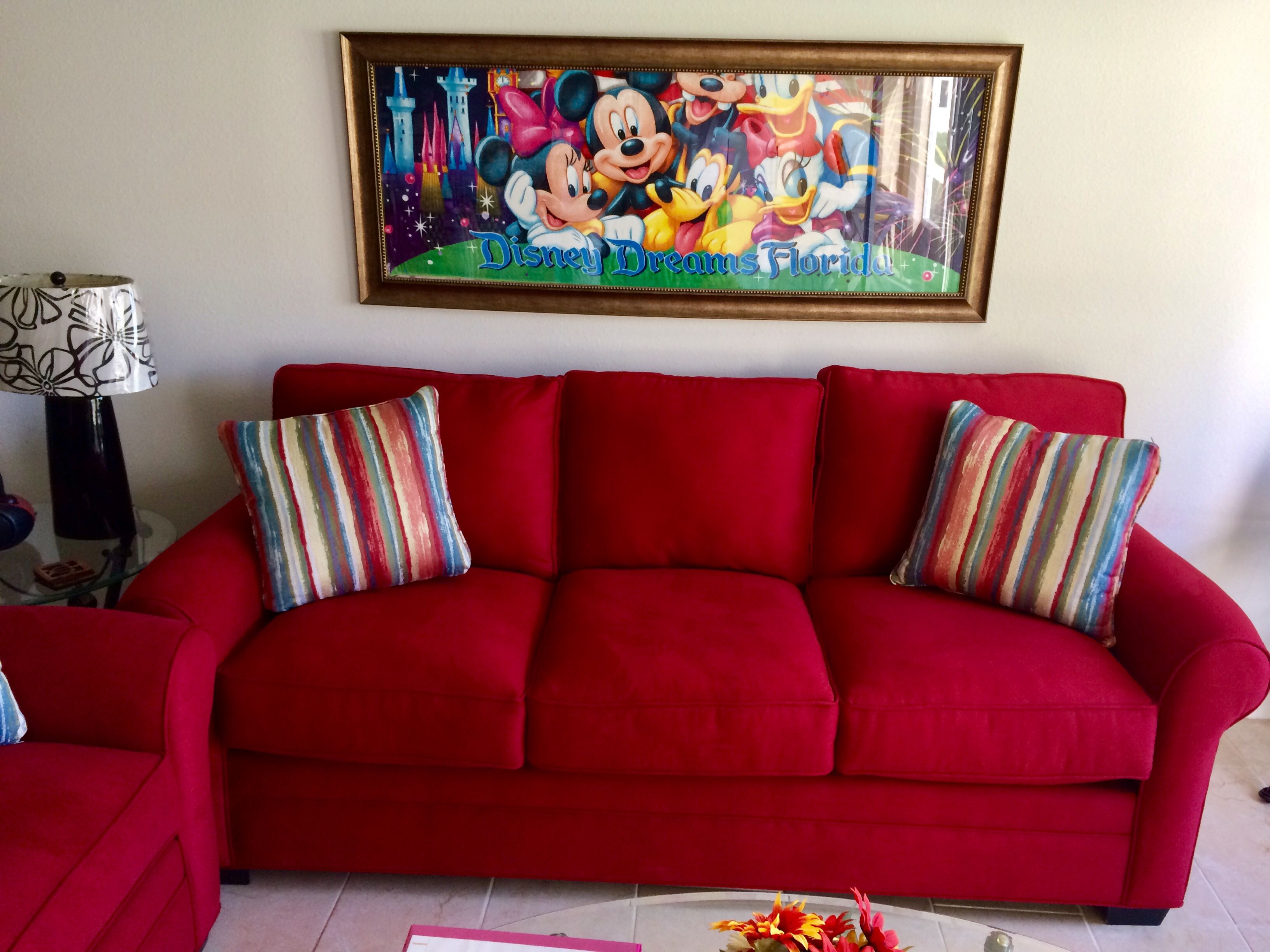 A red sofa with striped pillows, a framed Disney Dreams Florida poster, and a lamp with a patterned shade.