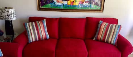Ein rotes Sofa mit gestreiften Kissen, ein gerahmter Poster von Disney Dreams Florida und eine Lampe mit gemustertem Lampenschirm.