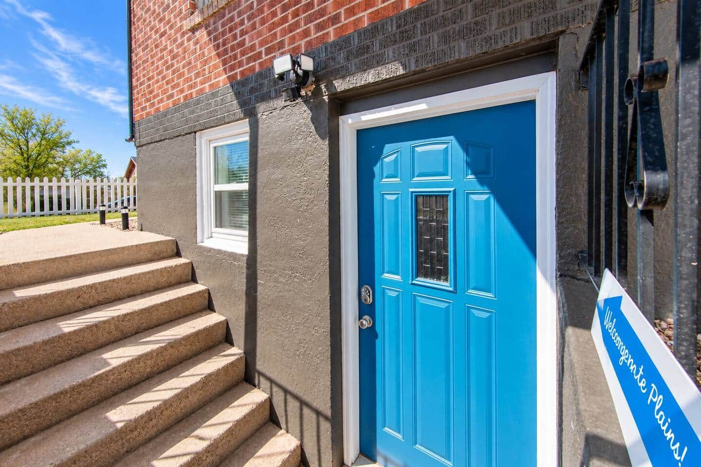 Une porte bleue avec une fenêtre, un escalier en béton menant à la porte, et une clôture blanche en bois en arrière-plan.