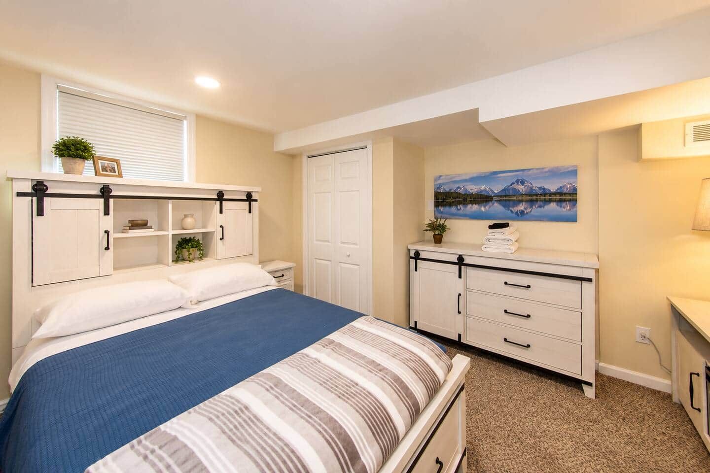 Een slaapkamer met een bed, een kaptafel, een nachtkastje, een raam met jaloezieën en een schilderij aan de muur.