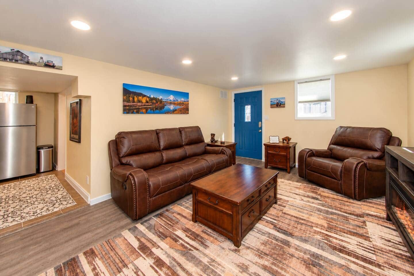 A living room with a brown leather sofa, a wooden coffee table, and two leather armchairs. There is a blue door, a refrigerator, and a painting on the wall.