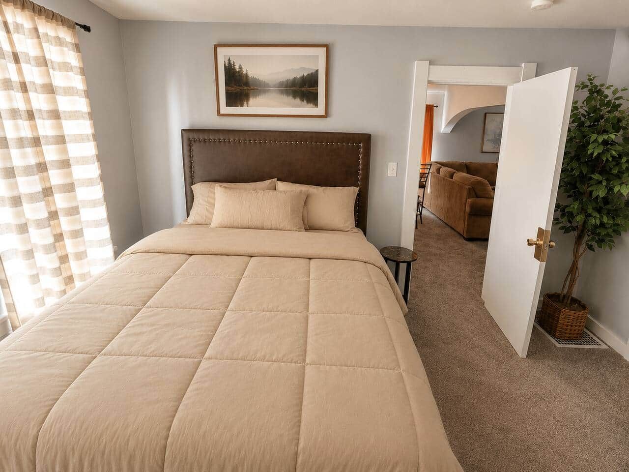 Une chambre à coucher avec un grand lit, une tête de lit, un tableau encadré au mur, une plante en pot et une porte donnant sur une autre pièce.