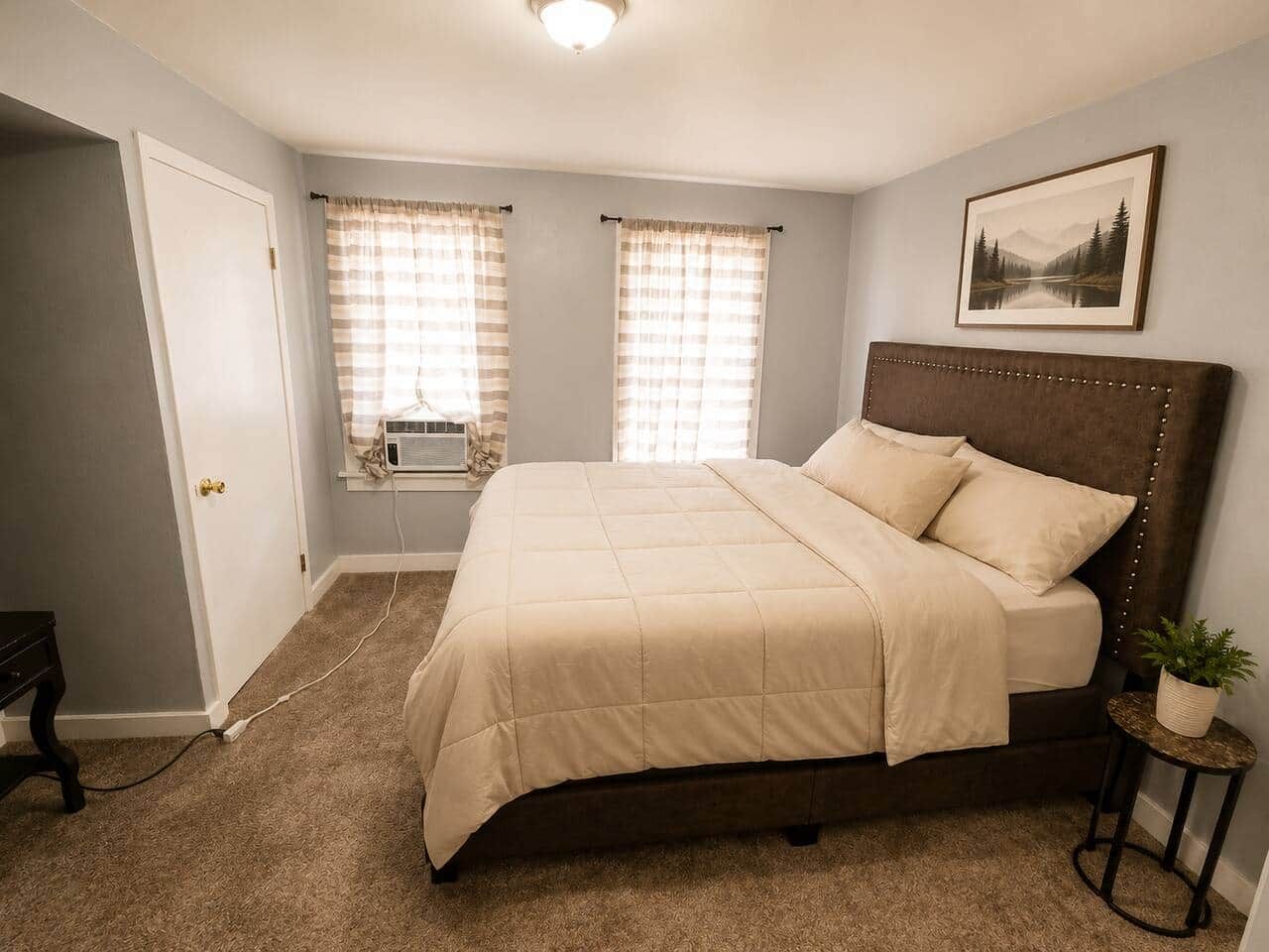 Une chambre à coucher avec un lit, une fenêtre avec des rideaux, un tableau accroché au mur et une petite table sur laquelle est posée une plante.